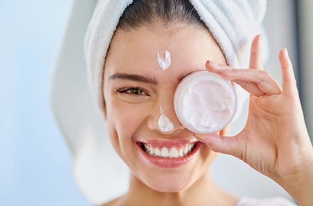 young_adult_woman_holds_facial_cream_to_right_eye_dabs_on_nose_smiles_towel_on_head_beauty