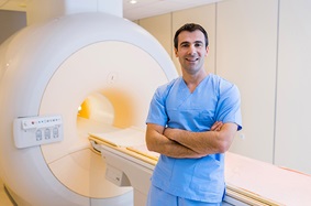 middle_aged_male_radiologist_wearing_blue_scrubs_stands_in_front_of_mri_machine