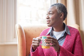 Older woman wearing a sweater while looking out the window and drinking coffee