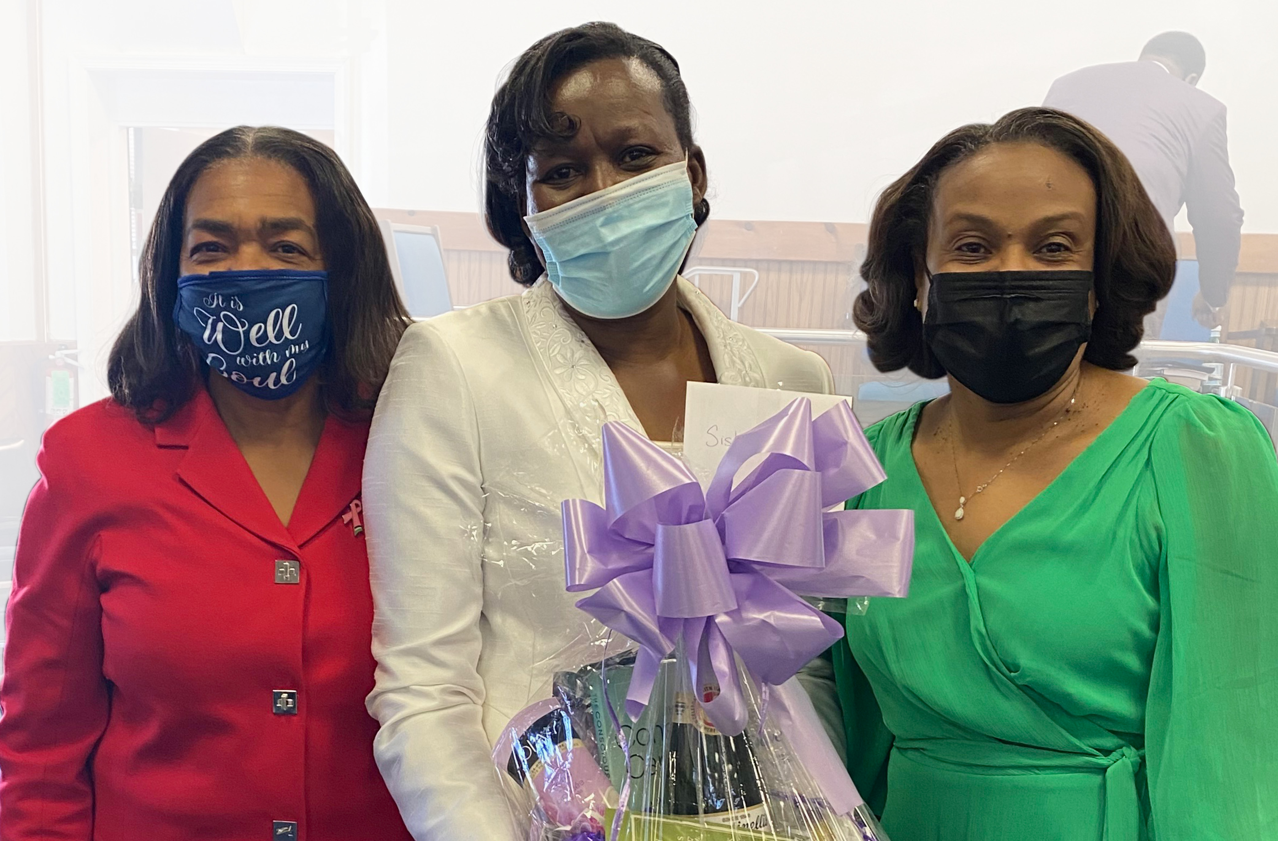 Three women in Sunday church dresses and face masks stand together. Left, Olive Campbell, coordinator of Caring for the Caregiver; Center, Neva Denton, a caregiver, holding a gift basket with a large purple ribbon; Right, Grace Hanson.