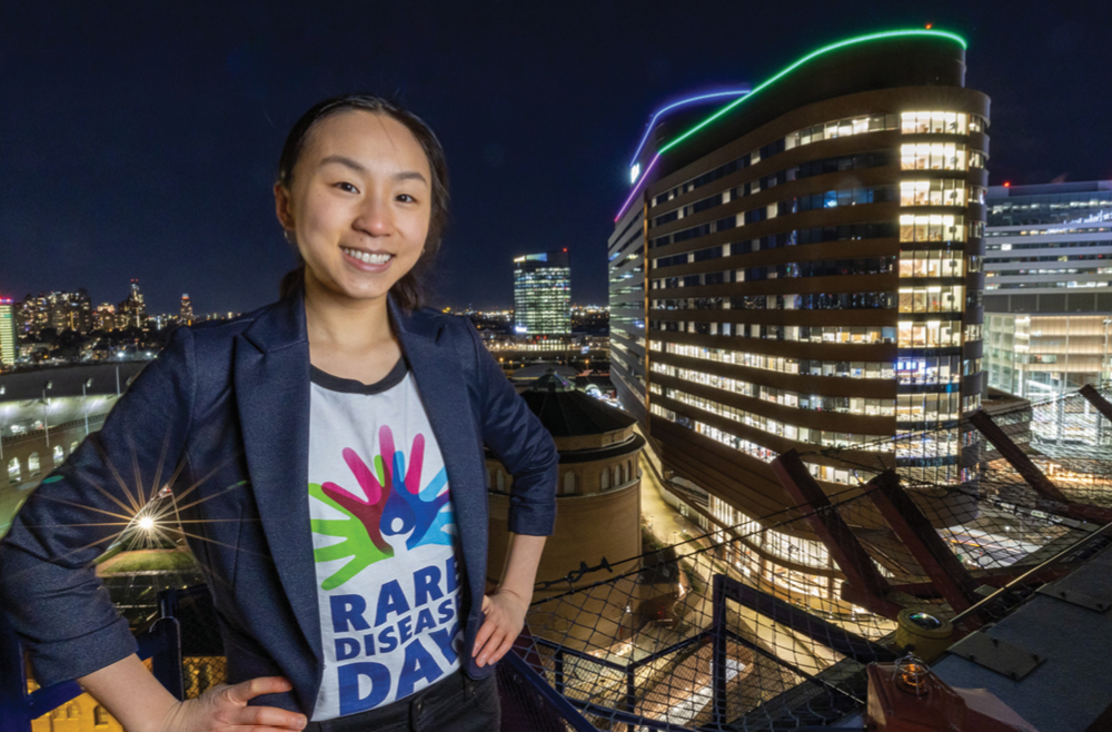 Magnolia Wang stands in front of the HUP Pavilion, lit up at night in blue, green, pink, and purple for Rare Disease Day.