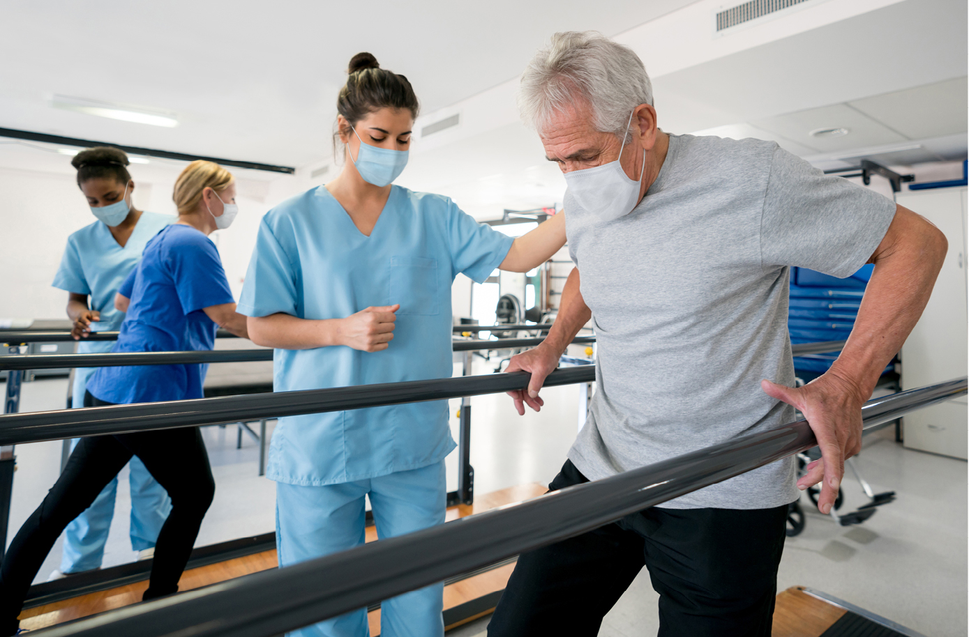 A man holds parallel bars for support during physical therapy. A therapist in scrubs stands nearby to spot him. Both wear facial masks.