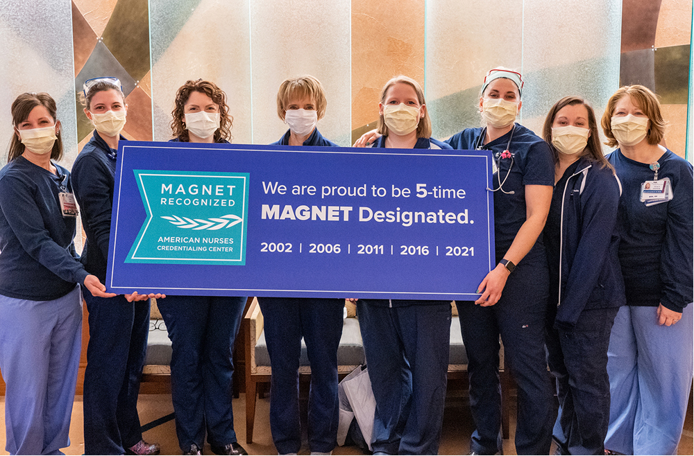 Eight nurses hold a large blue banner that reads: We are proud to be 5-time MAGNET Designated