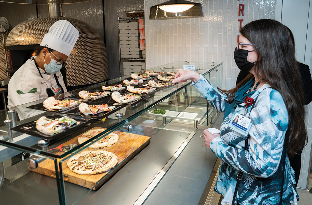 A chef prepares pizza behind a counter while an employee picks up a slice of pizza from atop the counter.